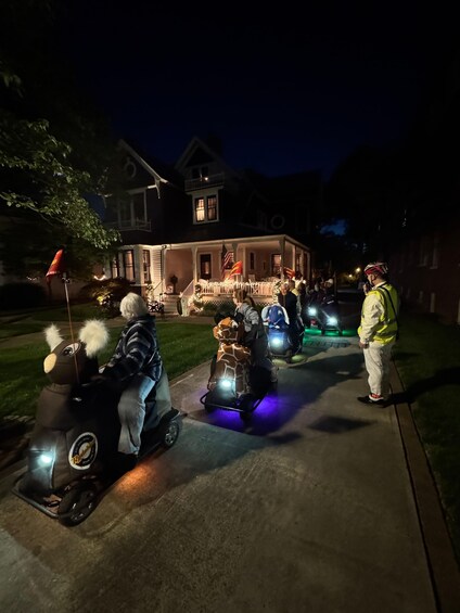 A nighttime scene with a house decorated with lights and flags, and people riding illuminated scooters. 4
