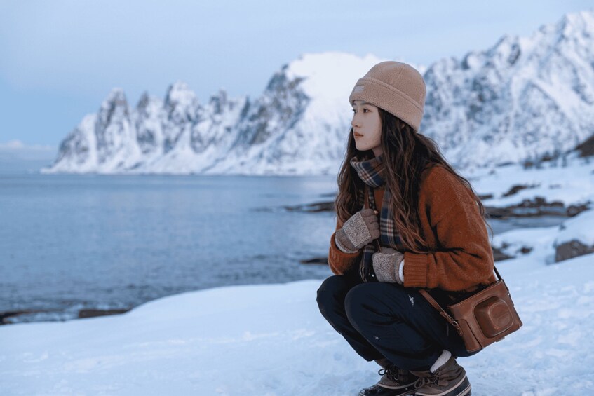 A person crouching on a snowy shore with a mountainous backdrop. 5