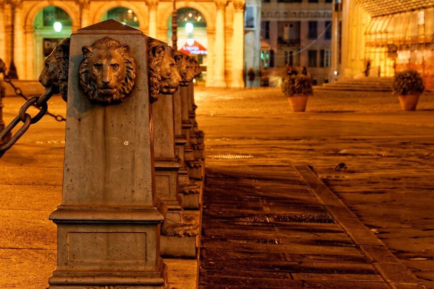 A row of lion head sculptures on a stone pedestal, with a cobblestone street and buildings in the background. 2