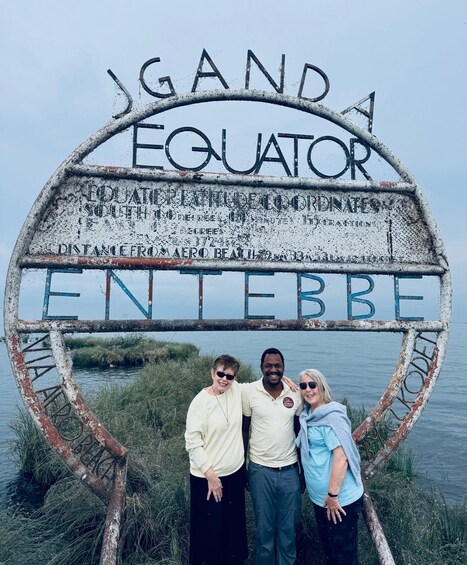 A weathered sign with 'Ganda Equator' and 'Entebbe' written on it, with three people standing in front. 1