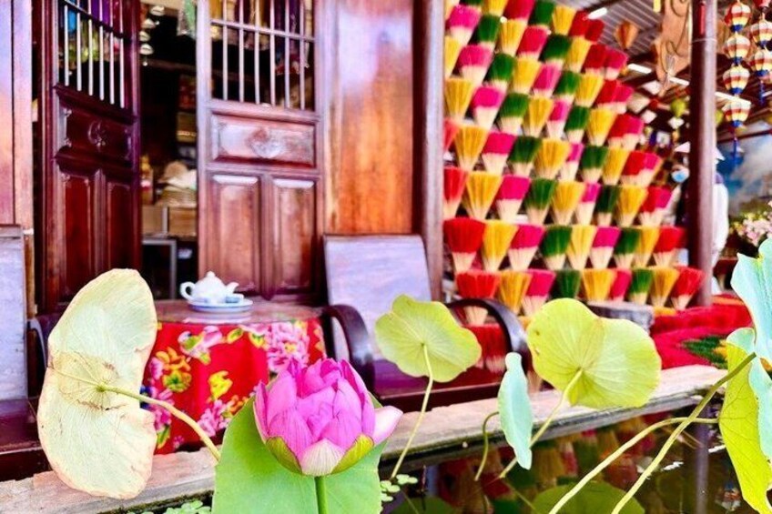 A traditional wooden house with a colorful quilt hanging on the wall, a table with a teapot, and lotus flowers in a pond. 5