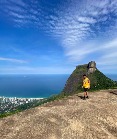 A person standing on a rocky outcrop overlooking a coastal city with a prominent mountain in the background. 2