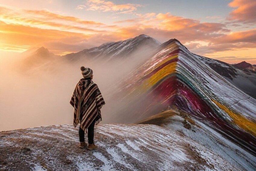 A person standing on a snowy mountain peak with a colorful, layered rock formation in the background. 3