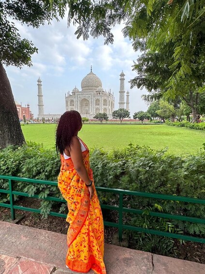 A person in an orange sari standing by a green railing, overlooking the Taj Mahal. 2