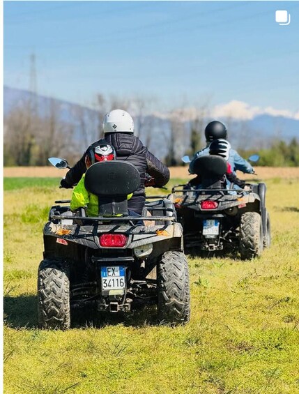 Two people riding ATVs in a grassy field with mountains in the background. 3