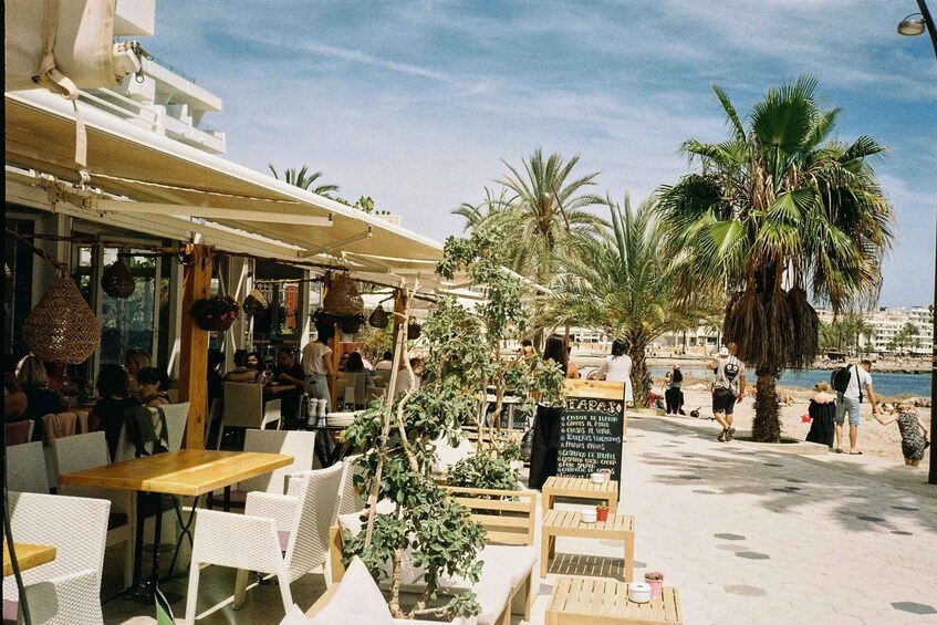 An outdoor café with tables and chairs, palm trees, and a view of the sea. 3