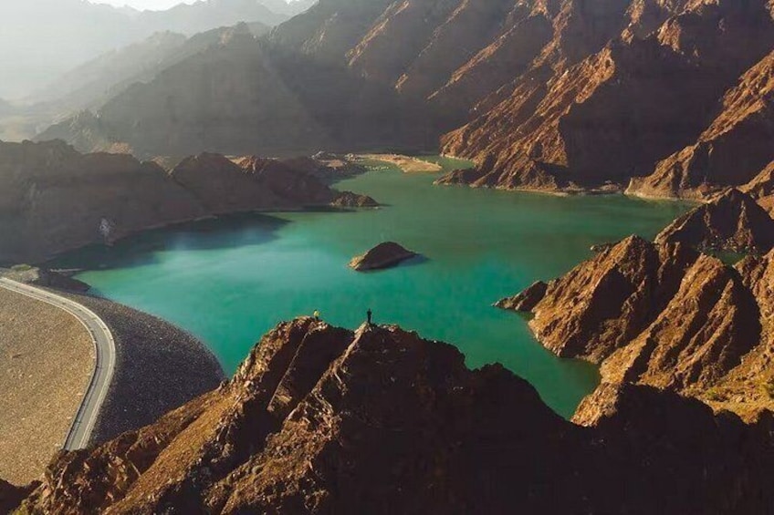 A mountainous landscape with a turquoise lake, a winding road, and a person standing on a hill. 5