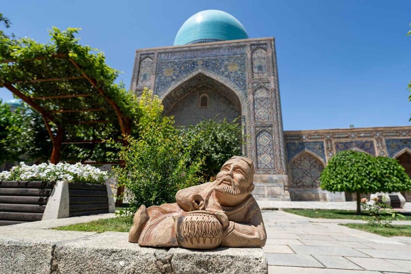 A stone statue of a seated figure with a bowl, in front of a building with a large dome and intricate tile work. 2