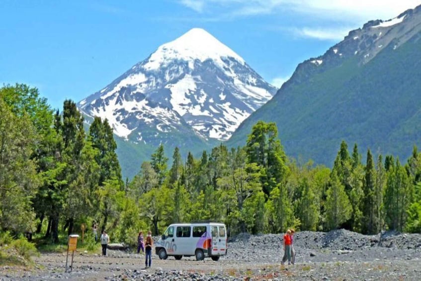 A white van parked in a rocky area with people around it, a snow-capped mountain in the background, and dense green trees. 1