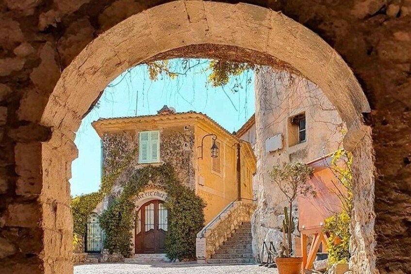 An arched stone doorway leading to a courtyard with a building featuring a tiled roof and a door with a sign above it. 1