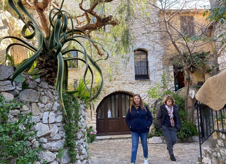 Two people walking in a stone courtyard with a large tree and a building with a wooden door. 5