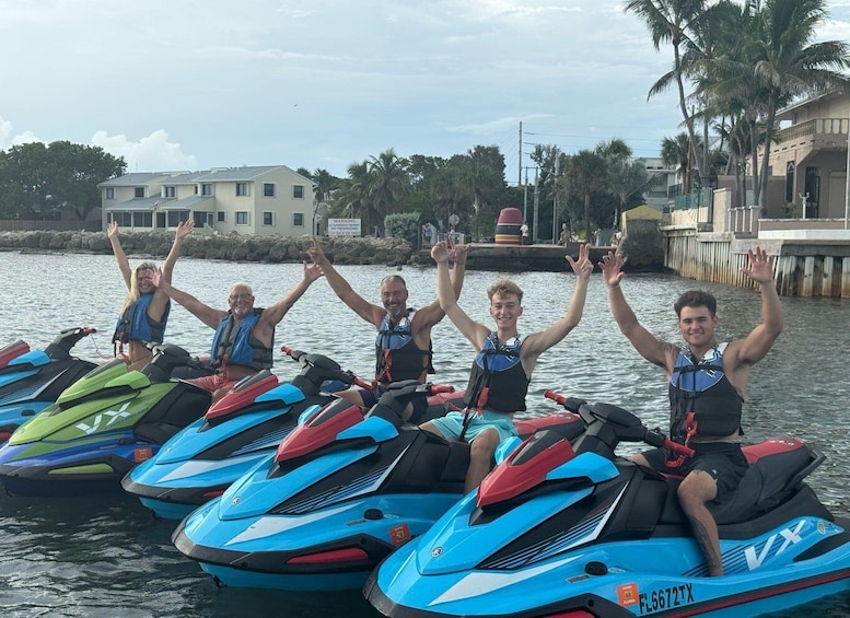 A group of people on jet skis, raising their hands in the air, with a waterfront and buildings in the background. 1