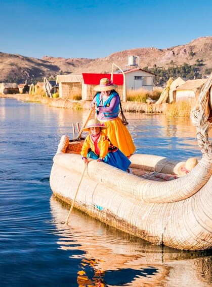 A person in traditional attire on a woven boat in a calm body of water, with a house and hills in the background. 1
