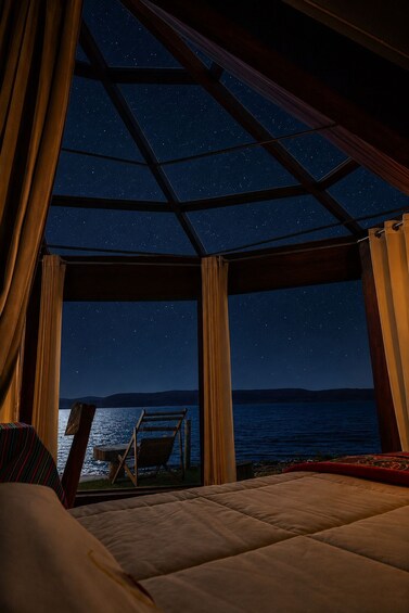 A bedroom with a view of a starry night sky, a bed, and a wooden deck overlooking a body of water. 3