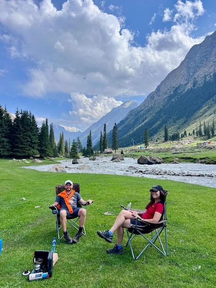 Two people sitting on lawn chairs in a grassy field with a river and mountains in the background. 1