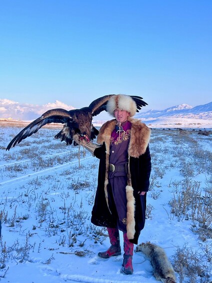 A person in traditional attire holding a large bird of prey in a snowy landscape with mountains in the background. 3