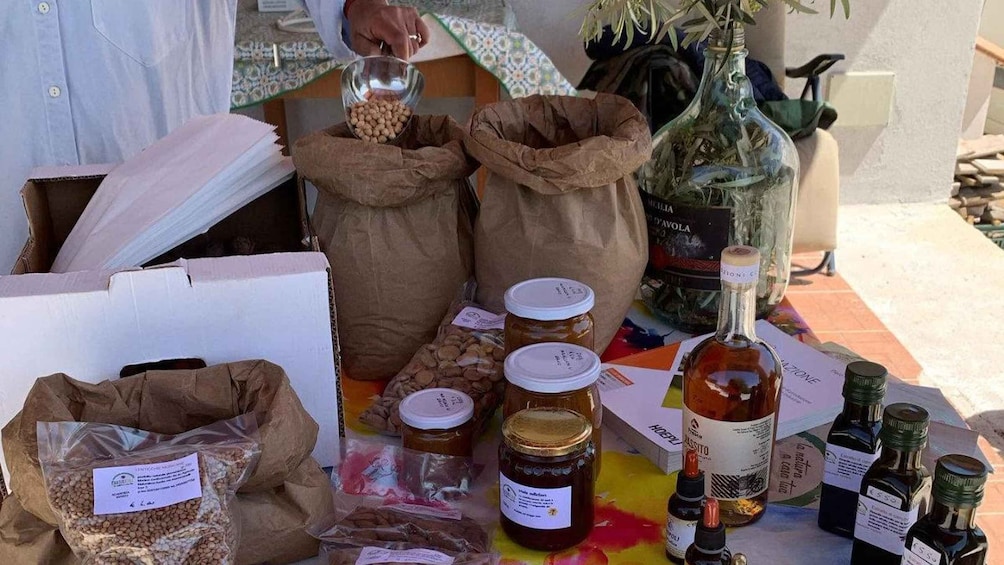 A table with various food items, including jars of preserves, bottles of olive oil, and bags of grains. 4