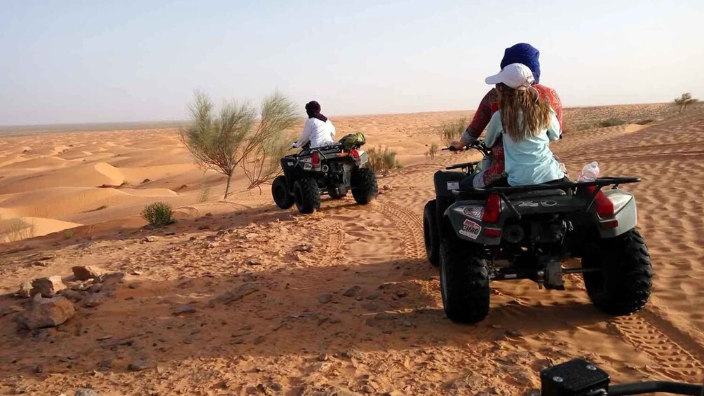 Two people riding ATVs in a desert with sand dunes and sparse vegetation. 3