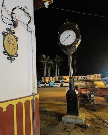 A street clock mounted on a pole, a sign reading 'Calle Real,' and parked cars and a motorcycle in the background. 4
