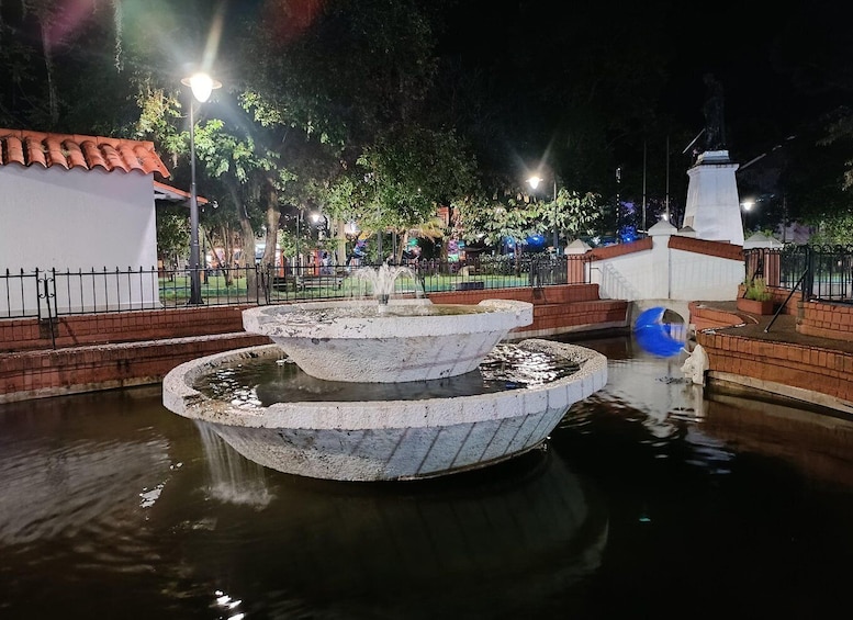 A nighttime fountain with water jets, surrounded by a brick wall and a white building with a red-tiled roof. 3