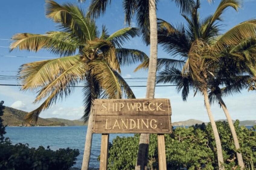 A wooden sign with the words 'SHIP WRECK LANDING' in front of palm trees and a body of water. 2