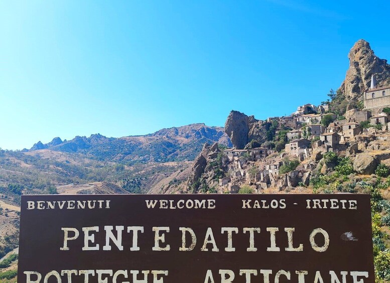 A sign with 'BENVENUTI WELCOME KALOS - IRIETE PENTEDATTILO' and 'ROTTETCHE ARTICIANE' indicating a welcoming sign in a mountainous area with traditional stone buildings. 4