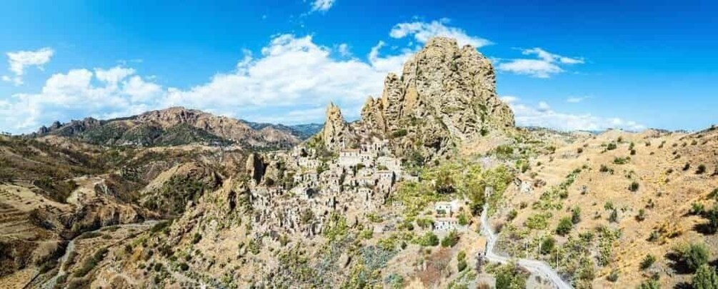 A rugged mountain landscape with a prominent rocky peak, a winding road, and scattered buildings. 3
