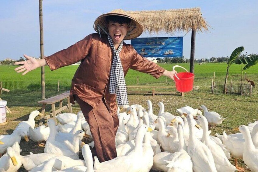 A person in a straw hat and brown outfit feeding ducks in a field with a thatched-roof structure in the background. 1