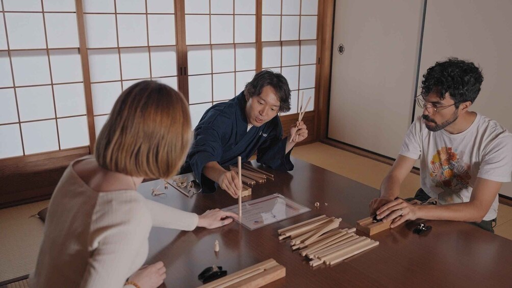 Three people sitting at a table with wooden sticks and a box. 3