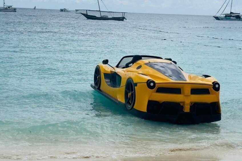 A yellow sports car parked on a beach with clear turquoise water and boats in the background. 5
