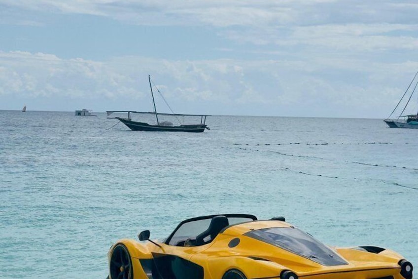 A yellow sports car on a beach with boats in the background. 4