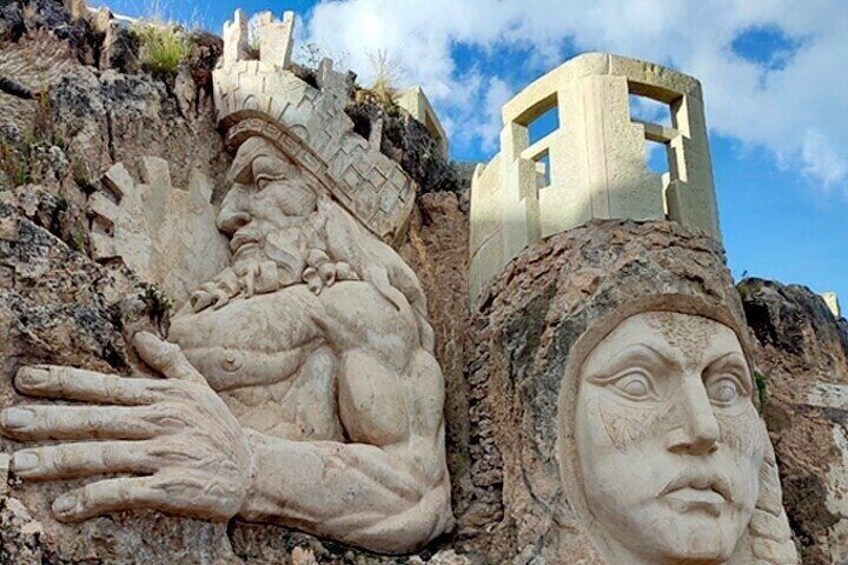 A large stone sculpture of a reclining figure with a hand extended, set against a rocky cliff with a small tower in the background. 4
