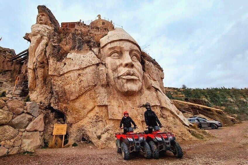 A large rock face with a carved face, a statue, and a dirt road with vehicles. 1