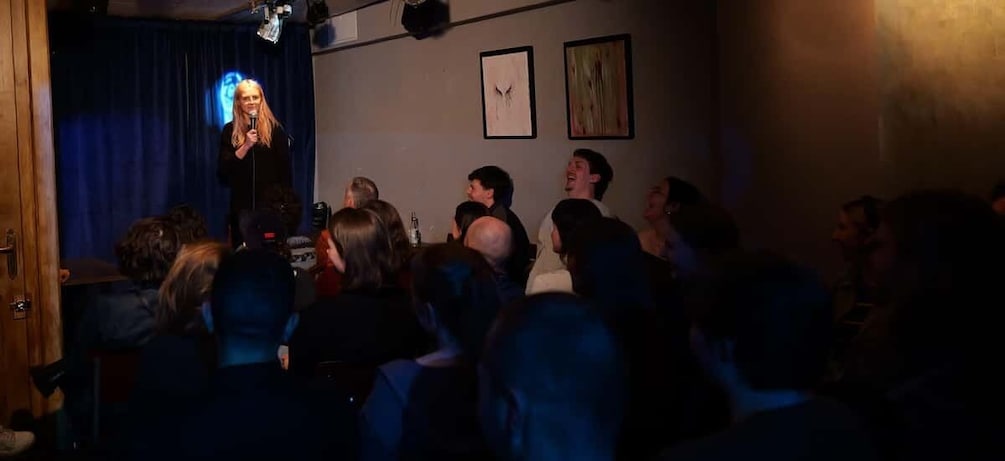 A person speaking to an audience in a dimly lit room with a blue curtain backdrop. 5