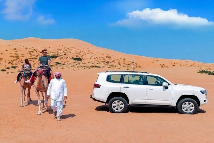 A desert landscape with sand dunes, a white SUV, and two camels with riders. 1