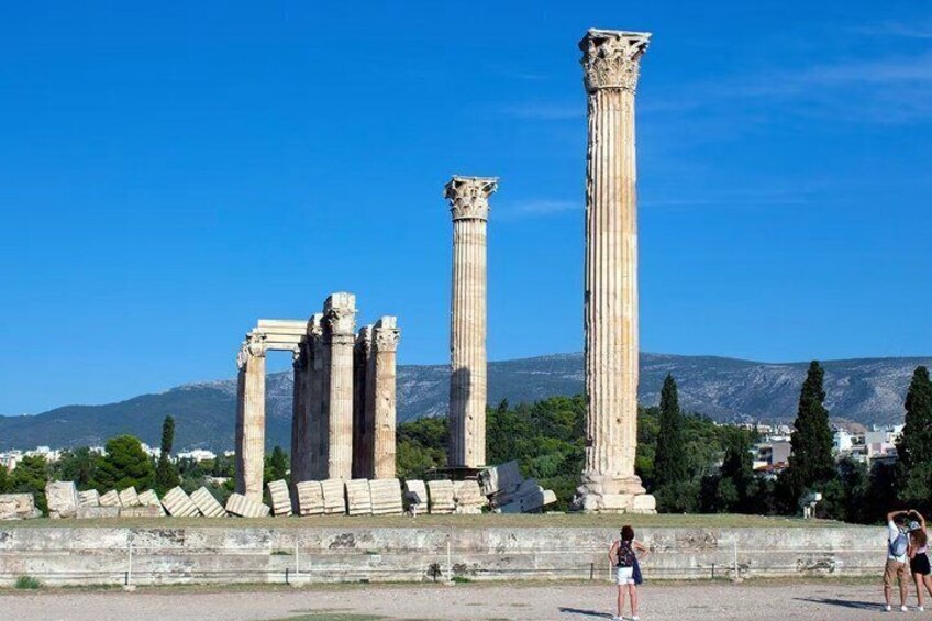 Ancient Greek columns, a clear sky, and a mountainous backdrop. 1