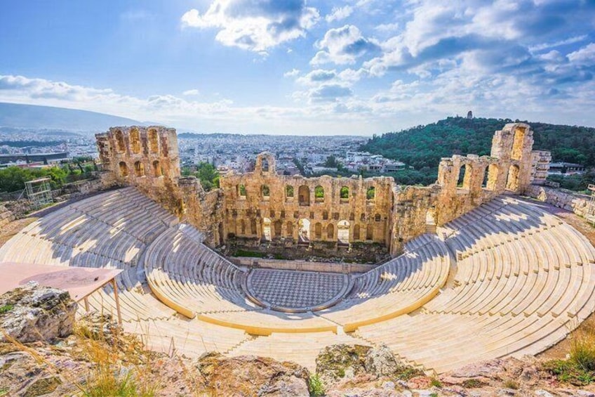 An ancient amphitheater with a circular stage and tiered seating, surrounded by ruins and a cityscape in the background. 3