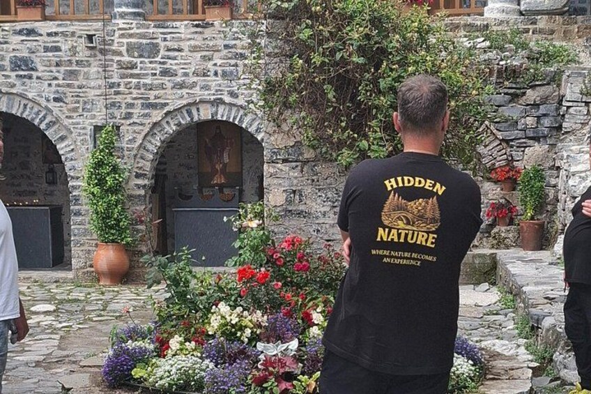 A man wearing a black t-shirt with 'HIDDEN NATURE' printed on the back, standing in front of a stone building with arched windows and a flower arrangement. 4