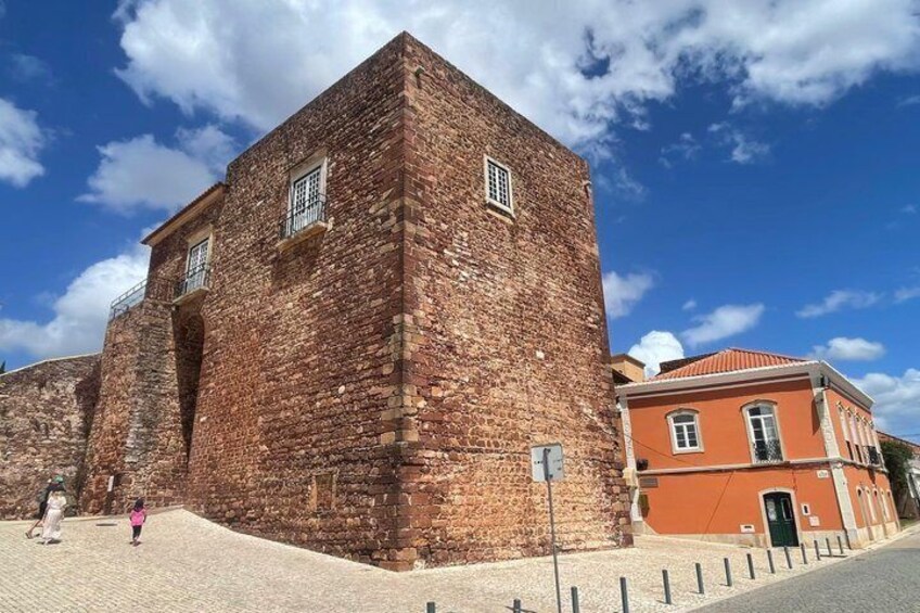 A historic brick building with a red-tiled roof, a paved area in front, and a clear blue sky. 4