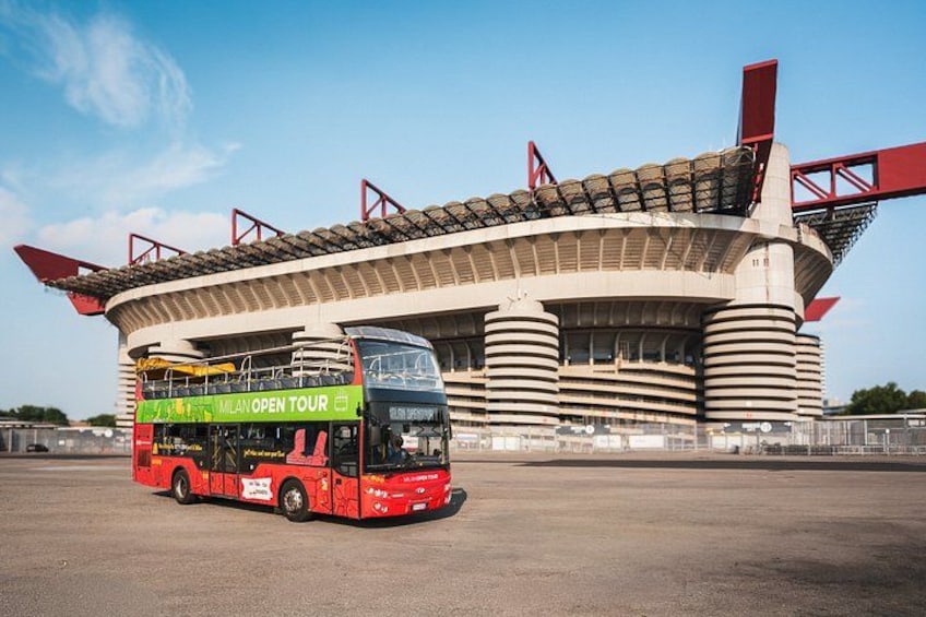 A red double-decker bus parked in front of a stadium with a green sign reading 'Milan Open Tour'. 2