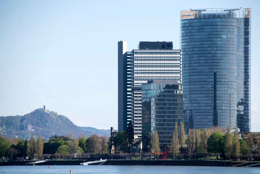 A cityscape with modern buildings, a river, and a hill with trees in the background. 1