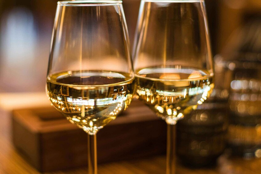 Two wine glasses filled with a clear liquid, likely wine, on a wooden surface with blurred background elements. 1