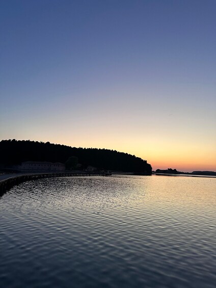 A calm body of water reflecting the sky during sunset, with a silhouette of a forested hill and a building in the background. 3