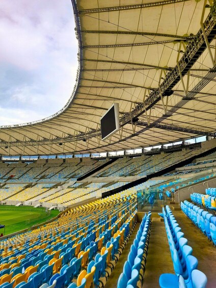 An empty stadium with blue and yellow seats, a large overhead structure, and a green field. 4