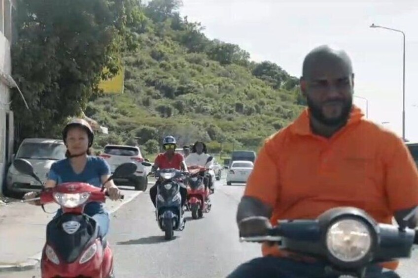A group of motorcyclists riding on a road with cars parked on the side, surrounded by greenery. 5