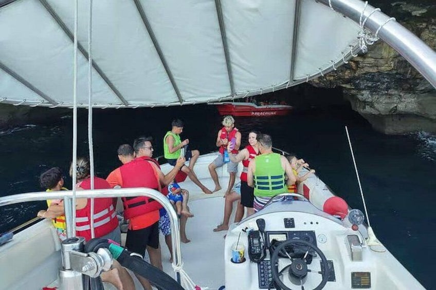 A group of people on a boat with a sail, some wearing life jackets, and a rocky shoreline in the background. 4