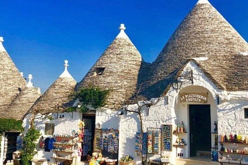 Traditional stone houses with conical roofs, a sign indicating a local shop, and a clear blue sky. 2