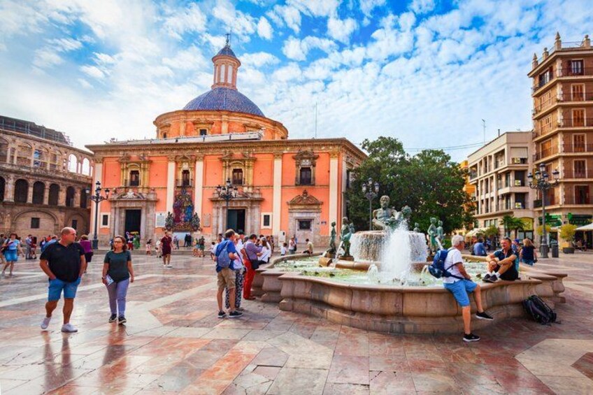 A public square with a fountain, a large building with a dome, and people walking around. 1