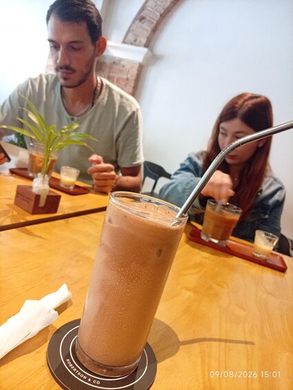 A person sitting at a wooden table with a glass of iced coffee, a napkin, and a small potted plant. 2