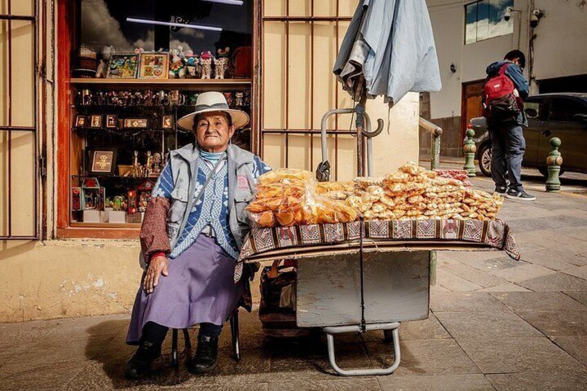 An elderly man sitting next to a cart filled with bread, with a shop in the background. 1
